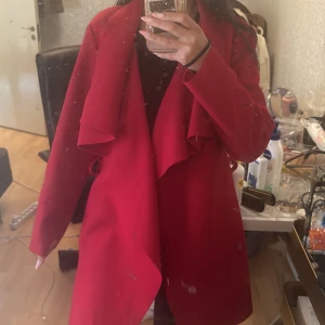 Röd omlottkappa med bred krage - Snygg röd kappa med omlottmodell och bred, draperad krage. Jackan har en stilren look och är perfekt för dig som vill sticka ut med färg. Passar till både jeans och klänning. Knytband i midjan för en snygg siluett.