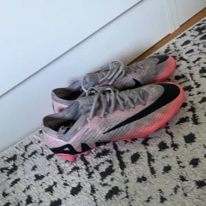 Nike Zoom rosa/grå fotbollsskor - Snygga Nike Zoom fotbollsskor i grått och rosa med svarta detaljer och stor Nike-logga på sidan. Skorna har snörning och plös i mesh, samt rosa dobbar för bra grepp på planen. Perfekta för dig som vill sticka ut på matchen!