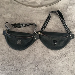 Svart magväska från Philipp Plein - Säljer en svart magväska från Philipp Plein med coolt mönster och detaljer. Väskan har ett präglat PP-mönster, silvrig dragkedja med rakbladshänge och bred axelrem med logga. Framsidan har en emblem med panterhuvud och en med PP 78. Perfekt för att lyfta din streetstyle. 200kr st