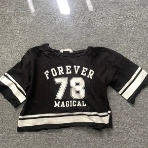 Svart croppad topp H&M Forever 78 - Cool svart croppad topp från H&M med vita tryck och texten 'FOREVER 78 MAGICAL' framtill. Tjocka vita ränder på ärmar och nederkant ger en sportig vibe. Perfekt till jeans eller shorts för en avslappnad look.