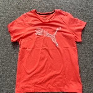 Rosa Puma t-shirt med logga XL - Snygg rosa t-shirt från Puma i storlek XL. Klassisk modell med rund hals och korta ärmar. Stor vit Puma-logga framtill. Tillverkad i mjuk bomull som känns skön mot huden. Perfekt för dig som gillar sportig och avslappnad stil. (Färgen på bilden ser inte ut som i verkligheten)