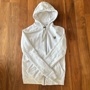 Vit hoodie från Lyle & Scott med dragkedja - Vit hoodie från Lyle & Scott med klassisk gul logga på bröstet. Tröjan har huva med snörning, hel dragkedja och två fickor framtill. Tillverkad i mjuk bomull och har långa ärmar. Perfekt för en clean och stilren look. Pris kan diskuteras. Och kan stryka plagget om du vill.