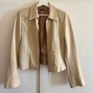 Vintage skinnjacka rock n blue - Snygg beige skinnjacka från rock n blue med klassisk krage och dragkedja framtill. Jackan har lång ärm med dragkedjedetalj vid ärmslut och en figurnära passform. Perfekt för dig som vill ha en clean och stilren look.