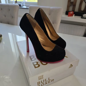 Svarta pumps från Bullboxer med silverklack - Stilrena svarta pumps från Bullboxer med höga silverfärgade klackar och rosa sula. Skorna har rund tå och är klädda i mocka för en lyxig känsla. Perfekta för dig som vill sticka ut med en färgklick under foten.