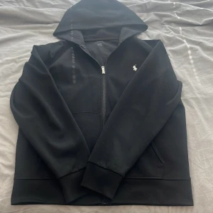 Svart hoodie från Polo Ralph Lauren - Svart hoodie från Polo Ralph Lauren med dragkedja och klassisk broderad logga på bröstet. Tröjan har huva, känguruficka och långa ärmar. Tillverkad i mjuk bomullsmix som är skön att bära. Perfekt för en chill och stilren look.