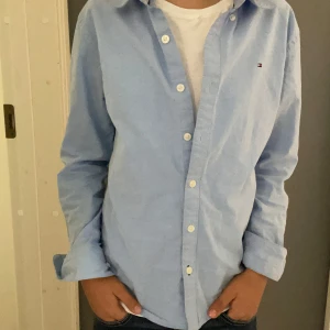 Ljusblå skjorta från Tommy Hilfiger - Snygg ljusblå skjorta från Tommy Hilfiger med klassisk krage och vita knappar. Skjortan har lång ärm och liten broderad logga på bröstet. Perfekt att bära öppen över en t-shirt eller knäppt för en clean look.