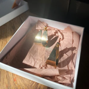 Guldiga statement örhängen - Säljer ett par coola guldiga hoops med bred, snirklig form. De har en modern och unik design som verkligen sticker ut. Perfekta för dig som vill ha något extra till din stil. Ej använda av mig, ifrån H&M