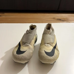 Nike Mercurial Air Zoom fotbollsskor - Snygga Nike Mercurial Air Zoom fotbollsskor i beige och svart med stora swoosh-loggor och textdetaljer. Skorna har en strumpliknande passform, kardborreband över vristen och plös för extra support. Ovandelen är i syntetmaterial och sulan har fasta dobbar för gräs.