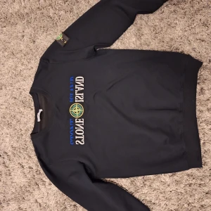 Svart Stone Island sweatshirt L - Svart sweatshirt från Stone Island med klassisk logga på ärmen och broderad text på bröstet. Tröjan har rund hals, ribbade muddar och är tillverkad i mjuk bomull. Perfekt för dig som gillar streetwear och vill ha en clean look.