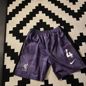 Lila Liverpool Nike shorts - Säljer ett par lila Liverpool FC shorts från Nike med vita detaljer och nummer 4 tryckt på ena benet. Shortsen har elastisk midja och ett mönster av tunna vita linjer över hela tyget. Perfekta för fotboll eller träning.