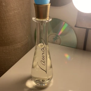 Laura Biagiotti Laura parfym - Elegant parfymflaska från Laura Biagiotti med genomskinligt glas, smal midja och bredare botten. Flaskans kork är guldig med en blå topp, och texten 'Laura' är tryckt i svart på framsidan. Perfekt för dig som gillar stilrena dofter.
