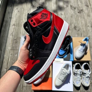 Nike Air Jordan 1 High Bred Satin - Nike Air Jordan 1 High i klassiska Bred-färger med svart och rött satinmaterial, vit sula och ikonisk röd Swoosh. Perfekt för dig som vill sticka ut lite extra med en ikonisk sneaker. Storlek 44,5 (US10,5/WMNS12) 