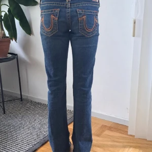 True Religion blå bootcut jeans - Säljer ett par klassiska blå jeans från True Religion med ikoniska kontrastsömmar och bakfickor med broderad logga. Modellen har bootcut-ben, låg midja och normal passform. Skriv gärna vid funderingar😊