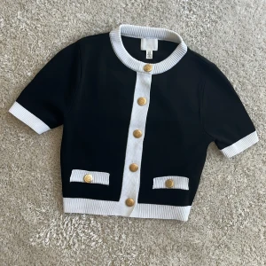 Svartvit kofta med guldknappar H&M XS - Stilren svart kofta från H&M i storlek XS med korta ärmar, vita ribbade kanter och dekorativa guldknappar framtill. Falska fickor med vita detaljer och guldknappar ger en lyxig känsla. Perfekt för dig som gillar klassisk och trendig stil. Använda endas ett fåtal gånger!!