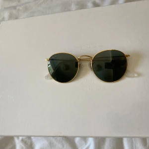 Ray-Ban runda solglasögon guld - Snygga Ray-Ban solglasögon med rund form och tunna guldfärgade metallbågar. Mörkgröna glas och klassisk design som ger en cool retrovibe. Näskuddar i transparent plast för extra komfort. Perfekta för dig som vill sticka ut med stil.