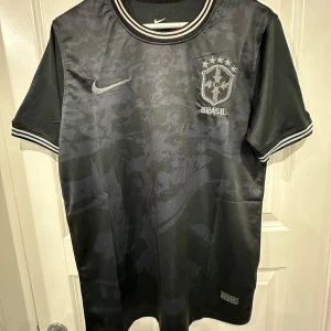 Svart Brasil Nike fotbollströja - Snygg svart Brasil fotbollströja från Nike med diskret camouflagemönster på framsidan. Broderat CBF-märke och Nike-logga i silver. Ribbad krage och ärmslut med vita och svarta ränder. Tillverkad i lätt och ventilerande material, perfekt för match eller träning.