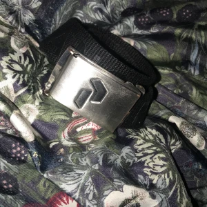 Svart tygbälte från Peak Performance - Säljer ett svart tygbälte från Peak Performance med en robust silverspänne i metall. Bältet har ett diskret tryck med märkesnamnet längs bandet och passar perfekt till jeans eller cargo. Stilren och enkel design som funkar till många outfits.