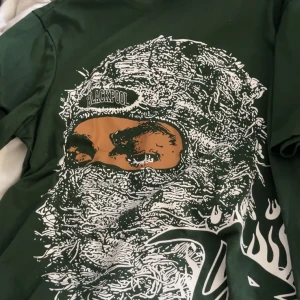 Grön t-shirt med grafiskt tryck från Romwe - Mörkgrön t-shirt från Romwe i storlek XS med stort grafiskt tryck framtill. Motivet visar ett ansikte med texten 'Blackpool' och coola vita flames. T-shirten har rund hals och korta ärmar, perfekt för dig som gillar streetstyle och unika prints.