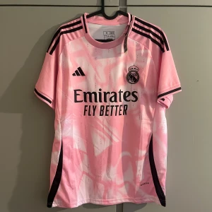 Rosa Real Madrid fotbollströja Adidas - Snygg rosa Real Madrid fotbollströja från Adidas med svart logga och klubbmärke på bröstet. Tröjan har korta ärmar, svarta ränder på axlarna och svart tryck framtill. Tillverkad i lätt och ventilerande material, perfekt för träning eller match.