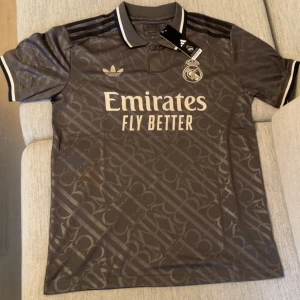 Real Madrid x Adidas fotbollströja grå - Snygg brun Real Madrid fotbollströja från Adidas med diskret mönster över hela tröjan. Klassisk krage med svarta och vita detaljer, korta ärmar och broderade loggor. Materialet är lätt och andas, perfekt för match eller träning. Emirates Fly Better tryck på bröstet.