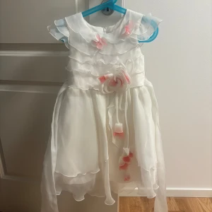 Vit kortklänning med volanger och blommor - Superfin vit kortklänning från NiGoBic med flera lager volanger och söta rosa blommor framtill. Klänningen har knytband i midjan och är ärmlös med romantiska detaljer. Perfekt för speciella tillfällen när du vill känna dig extra söt.
