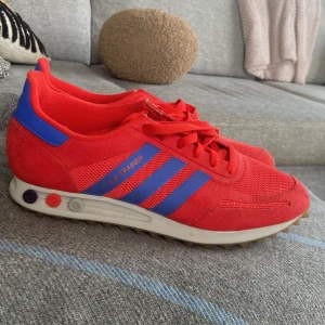 Adidas LA Trainer röda sneakers strl 44 - Snygga Adidas LA Trainer sneakers i rött med blå ränder och detaljer. Ovandel i mesh och mocka, klassisk retrostil med snörning och platt sula. Perfekta för dig som gillar färgstarka och sportiga skor.