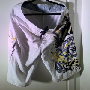 Quiksilver vita badshorts med tryck storlek 36.  - Snygga vita badshorts från Quiksilver med svart och gult grafiskt tryck, coola detaljer och snörning i midjan. Shortsen har en ficka på sidan med broderad logga och är perfekta för stranden eller poolen. Lätta och snabbtorkande material.