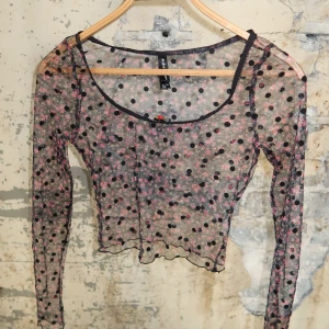 Mesh topp med prickigt och blommigt mönster - Säljer en transparent mesh topp från for love & lemons med svarta prickar och små rosa blommor. Toppen är croppad, har lång ärm och en söt liten rosett vid urringningen. Perfekt för att skapa en cool lager-på-lager look.