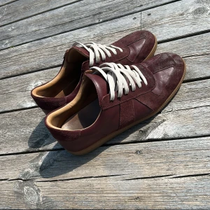 Vinröda sneakers i skinn och mocka - Säljer ett par vinröda GAT-sneakers med ovandel i både skinn och mocka, vita snören och klassisk gummisula. Skorna har en stilren design med rund tå och beige insida. Perfekta för dig som gillar retrovibbar och vill sticka ut med en djup färgton. Det ända är slitaget på färgen (finns en bild) annars är det inget fel på dom.