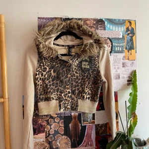 Leopardmönstrad croppad jacka - Säljer en croppad jacka med leopardmönster framtill, beige ärmar och huva med fluffig fuskpäls. Guldiga knappar och broderad CL-logga på bröstet. Jackan har ribbade muddar och cool streetstil. Tyvärr lite lien vid bicep för mig ! 