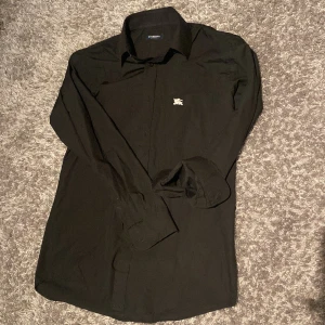 Svart skjorta från Burberry London - Stilren svart skjorta från Burberry London med klassisk krage och diskret broderad logga på bröstet. Skjortan har långa ärmar och knäppning framtill. Perfekt för dig som gillar en clean och tidlös look.