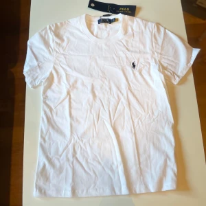 Vit t-shirt från Polo Ralph Lauren - Klassisk vit t-shirt från Polo Ralph Lauren i mjuk bomull. T-shirten har korta ärmar, rund halsringning och den ikoniska broderade loggan i svart på bröstet. Perfekt basic-plagg med clean look och snygg passform.