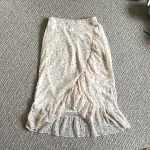 Blommig omlottkjol från Vero Moda - Säljer en långkjol från Vero Moda i ljusbeige med små rosa blommor. Kjolen har omlottmodell med volangkant nertill och resår i midjan. 