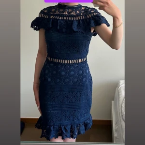 Marinblå spetsklänning med volang - Jättefin marinblå kortklänning i spets med volangdetaljer och genombrutna partier vid midjan och axlarna. Klänningen är ärmlös, med snygg volangkant nertill. 