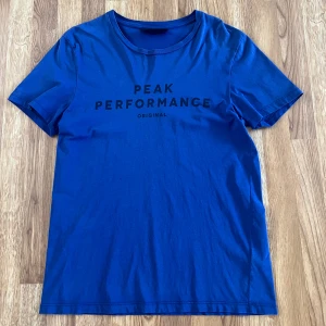 Blå Peak Performance t-shirt - Snygg blå t-shirt från Peak Performance med svart tryck på bröstet. Klassisk rund halsringning och korta ärmar. Tillverkad i mjuk bomull som känns skön mot huden. Perfekt för en avslappnad och sportig look.