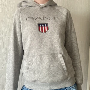 Grå hoodie från GANT med logga - Snygg ljusgrå hoodie från GANT med broderad logga och sköld på bröstet. Tröjan har huva, känguruficka och är tillverkad i mjuk bomullsmix. Perfekt för en chill och sportig look.