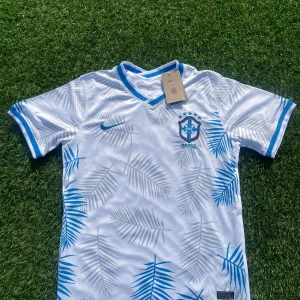 Vit Brasilien fotbollströja Nike L - Snygg vit Brasilien fotbollströja från Nike med blå detaljer och palmbladsmönster i grått och blått. Tröjan har korta ärmar, V-ringad krage med blå kant och är tillverkad i Dri-FIT-material som andas. Perfekt för dig som vill sticka ut på planen eller läktaren.