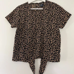 Leopardmönstrad topp från Seaside - Cool kortärmad topp från Seaside med leopardmönster i svart och beige. Toppen har rund hals och knytning framtill som ger en extra snygg detalj. Perfekt för dig som vill sticka ut med ett djurmönstrat plagg.