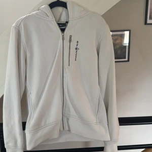 Vit hoodie från Sail Racing - Vit hoodie från Sail Racing med dragkedja framtill och en extra bröstficka med dragkedja. Tröjan har huva, långa ärmar och en clean look med logga på bröstet. Perfekt för dig som gillar stilren och sportig design. Använd sparsamt