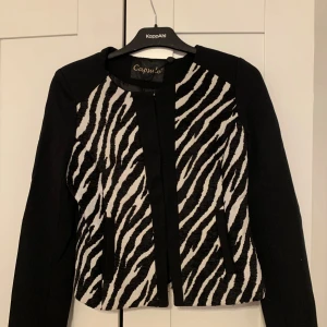 Zebramönstrad kavaj och kjol Capsule S - Snyggt set från Capsule med kavaj och kjol i svart och vitt zebramönster. Kavajen har raka ärmar och två fickor framtill, kjolen är figurnära och har samma coola zebraprint framtill. Perfekt för dig som vill sticka ut med en trendig look. Helt nya med prislappar kvar.