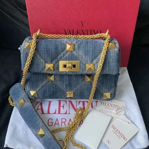 Valentino Garavani jeans axelväska - Snygg axelväska från Valentino Garavani i blått jeansmaterial med quiltad design och guldiga nitar. Väskan har kedjerem i guld och ett elegant vridlås framtill. Perfekt för dig som vill sticka ut med en cool och lyxig accessoar.