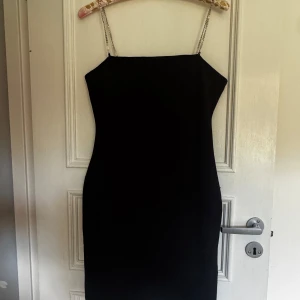 Svart kort klänning med strassband - Supersnygg svart kort klänning med raka linjer och smala axelband i strass som ger extra bling. Enkel och stilren design, perfekt för att sticka ut. Axelbandslös känsla men med glittriga band som detalj. Figurnära passform. Kan mötas upp i Norrköping 😊