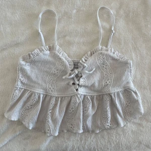 Vit croppad linnetopp från FB Sister - Säljer en söt vit croppad linnetopp från FB Sister med spetsdetaljer och volang. Toppen har smala axelband, knytning framtill och ett romantiskt mönster. Perfekt för varma dagar och sommarens alla tillfällen! Den är ganska nopprig