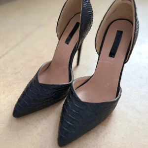 Svarta pumps med ormskinnsmönster - Säljer ett par svarta pumps med spetsig tå och hög klack. Skorna har ett elegant ormskinnsmönster och öppen insida, vilket ger en stilren och trendig look. Perfekta för dig som vill sticka ut med en klassisk men edgy stil. Helt nya, aldrig använt, bara testat