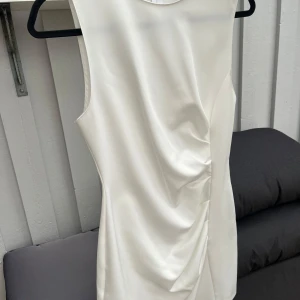 Vit omlottklänning från Zara - Säljer en elegant vit omlottklänning från Zara i storlek S. Klänningen är ärmlös med rund halsringning och har snygga draperade detaljer framtill. Tillverkad i ett slätt, glansigt material som ger en lyxig känsla.