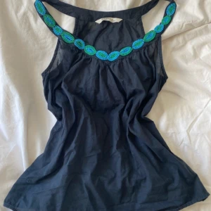 Svart linnetopp med pärldetaljer från Old Navy - Säljer en svart linnetopp från Old Navy med snygga blå och gröna pärldetaljer längs halsringningen. Toppen är ärmlös och har en stilren, enkel design som passar perfekt till sommaren.