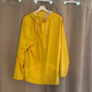 Gul regnjacka från Rains - Snygg gul regnjacka från Rains med huva och dragsko. Jackan har tryckknappar framtill, två fickor med lock och en rak passform. Perfekt för regniga dagar och enkel att matcha med andra plagg.