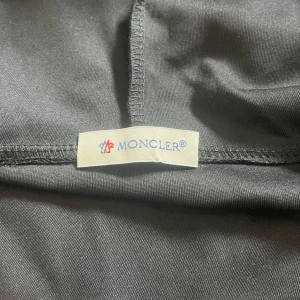 Svart hoodie med dragkedja från Moncler - Säljer en svart hoodie från Moncler med dragkedja och huva. Tröjan har vita och röda detaljer på ärmsluten och huvan samt Moncler-logga på ärmen. Perfekt för dig som gillar stilrena märkesplagg.