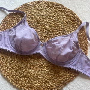 Ljuslila bh från Fenty - Säljer en ljuslila bh från Fenty med diskret logotypmönster i tyget och en liten guldfärgad detalj framtill. Bh:n har tunna axelband och är tillverkad i ett mjukt, syntetiskt material. Perfekt för dig som gillar stilrena underkläder med en twist.