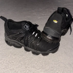 Nike Air Vapormax Plus Svarta Sneakers - Säljer ett par svarta Nike Air Vapormax Plus sneakers med klassisk bubbelsula och snörning. Skorna har en sportig design med diskreta Nike-loggor och detaljer i syntetmaterial. Perfekta för dig som gillar streetwear och vill ha något bekvämt och stilrent.